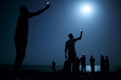 African migrants on the shore of Djibouti City at night raise their phones in an attempt to catch an inexpensive signal from neighboring Somalia—a tenuous link to relatives abroad. Djibouti is a common stop-off point for migrants in transit from such countries as Somalia, Ethiopia and Eritrea, seeking a better life in Europe and the Middle East. (Photo by John Stanmeyer) - Source: World Press Photo